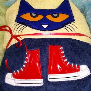 May include: Blue and orange cat face embroidered on a yellow hooded towel with red ribbon ties. The towel has red and white embroidered sneakers on the bottom.