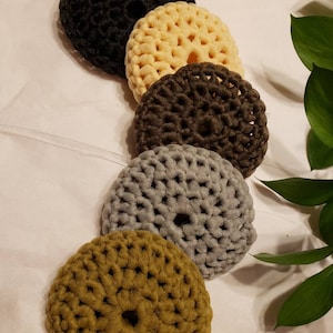 May include: Five crocheted hair scrunchies in different colors: black, cream, brown, gray, and olive green. The scrunchies are arranged in a row from left to right.