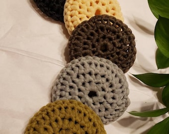 May include: Five crocheted hair scrunchies in different colors: black, cream, brown, gray, and olive green. The scrunchies are arranged in a row from left to right.