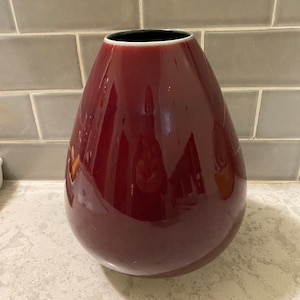 May include: A deep red ceramic vase with a rounded, pear-shaped body. The vase has a glossy finish and a dark rim.