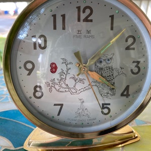 May include: A gold-colored alarm clock with a white face and black numbers. The clock has a picture of an owl on a branch with the words "Five Rams" and "Guangzhou China" printed on the face.