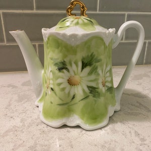 May include: A white porcelain teapot with a green and white floral design. The teapot has a gold lid handle and a scalloped edge.