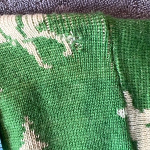 May include: Close-up of a green and white knitted sweater with a pattern of trees. The sweater is made of a soft, comfortable material.