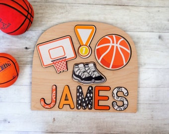 Puede incluir: Un rompecabezas de madera con temática de baloncesto con el nombre "JAMES" escrito con letras coloridas. El rompecabezas incluye imágenes relacionadas con el baloncesto como una canasta, una pelota, zapatillas y una medalla. Dos balones de baloncesto son visibles en el fondo.