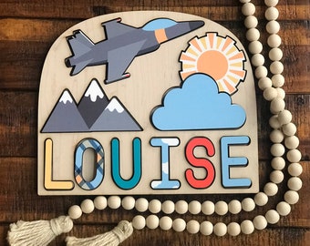 May include: A wooden puzzle shaped like an arch with a blue cloud, a yellow sun, a gray mountain range, a gray and blue airplane, and the name "LOUISE" in colorful letters.