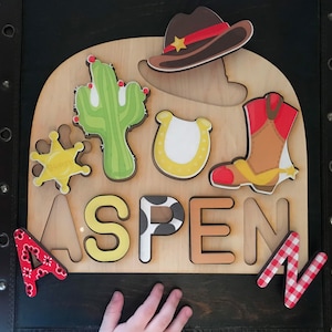 May include: Wooden puzzle with a cowboy theme. The puzzle pieces include a cactus, a horseshoe, a cowboy boot, a sheriff's badge, and the letters A, S, P, E, and N. The puzzle is on a brown wooden background.