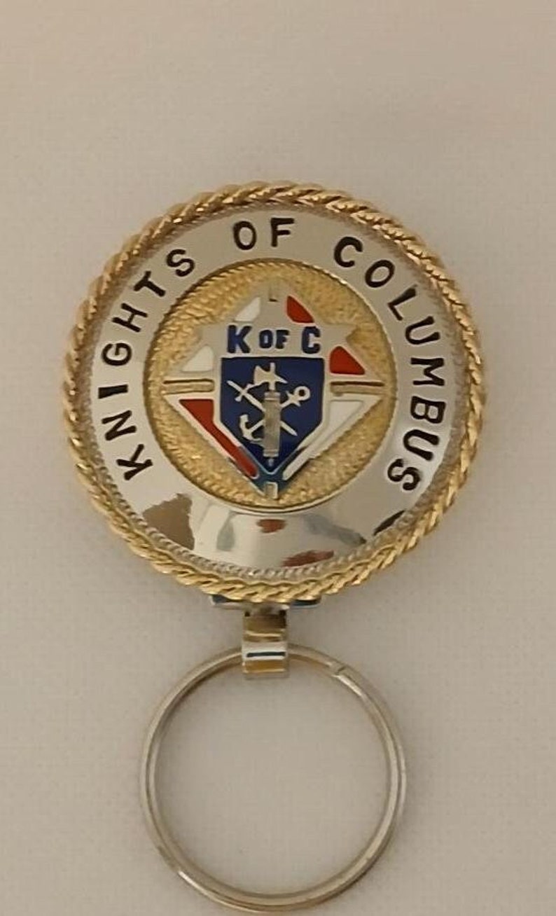 Puede incluir: Un llavero de oro y plata con una etiqueta redonda con borde de cuerda que dice "Knights of Columbus" y presenta un emblema azul, blanco y rojo con las letras "K of C".