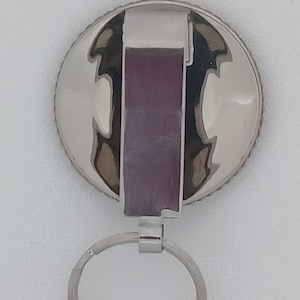 Puede incluir: Llavero plateado con un dije redondo de metal plateado y un anillo plateado. El dije tiene una superficie texturizada y estampada.