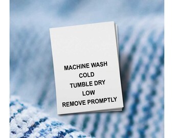 May include: A white clothing care label with black text. The label reads "MACHINE WASH COLD TUMBLE DRY LOW REMOVE PROMPTLY". The label is slightly angled, with a blurred blue and white background.