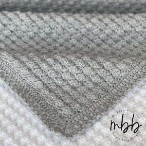 May include: A close-up of a gray crocheted blanket with a textured, raised pattern. The blanket is folded over, revealing a corner. The blanket is made of soft yarn and is suitable for a baby or child.