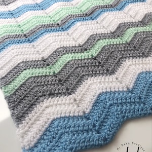 Op de afbeelding: Een gehaakte babydeken met een chevron-patroon in tinten blauw, groen, grijs en wit. De deken is gemaakt met een eenvoudige steek en heeft een gegolfde rand. De deken is gelabeld met "My Baby Boutique" en "mbb Simple Baby Designs".