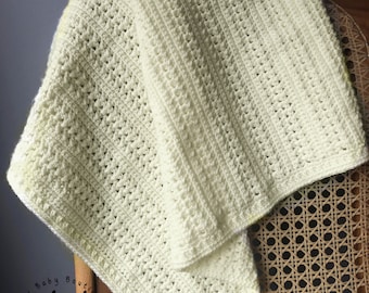 May include: A crocheted baby blanket in a light green and white color scheme. The blanket is folded and draped over a wicker chair. The blanket has a white border.