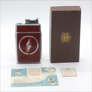 May include: A vintage red and silver lighter with a geometric design, a brown box with a gold emblem, and two small cards. The lighter has a rectangular shape with a circular design on the front. The box is rectangular and has a hinged lid.