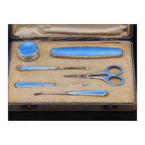 May include: A blue enamel manicure set in a velvet-lined case. The set includes a nail file, scissors, tweezers, a cuticle pusher, and a small jar.