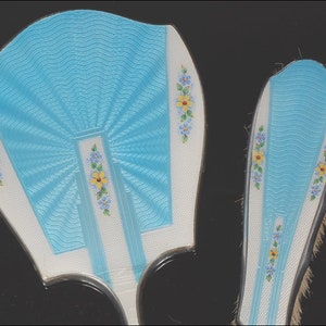 May include: A vintage vanity set featuring a hand mirror and a brush. The set has a light blue, fan-shaped design with white accents and floral details. The brush has a similar design with bristles.