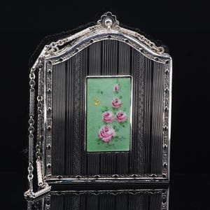 May include: A rectangular silver compact case with a pointed arch top and a delicate silver chain. The surface features black vertical stripes and a central rectangular panel in light green, decorated with a painted cluster of pink roses.