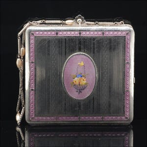 May include: A silver cigarette case with a decorative floral design. The case features a rectangular shape with a textured surface and a pink and purple border. An oval-shaped inset displays a basket of flowers.