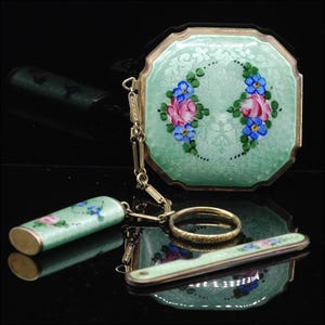May include: A vintage green enamel compact with a floral design and a matching lipstick holder and a small knife. The compact has a chain attached to it.