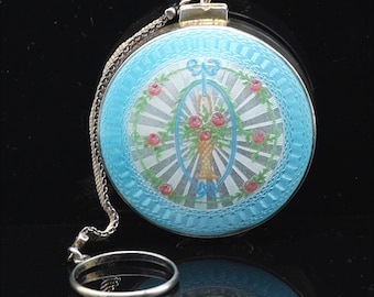 May include: A round, silver compact with a blue enamel background and a floral design. The compact has a chain attached to it.
