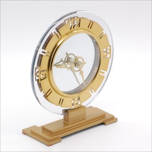May include: A circular table clock featuring a clear acrylic frame with a gold-toned metallic ring. The face displays gold-colored numerals at the three, six, nine, and twelve o'clock positions, with matching gold hands mounted on a rectangular base.