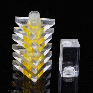 May include: A clear, geometric perfume bottle with a pyramid design, featuring alternating layers of clear and yellow acrylic. The bottle has a clear plastic neck and a separate, clear rectangular stopper. The design is modern and minimalist.