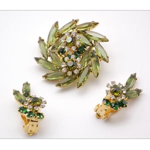 Puede incluir: Un conjunto de broche y pendientes en tono verde y dorado con diamantes de imitación transparentes y verdes. El broche tiene un diseño de remolino y los pendientes tienen un diseño de racimo.