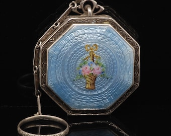 May include: An antique silver octagonal compact with a light blue enamel surface. The compact features a floral basket design in the center. A small silver ring is attached to the compact by a chain. The compact is a vintage accessory.