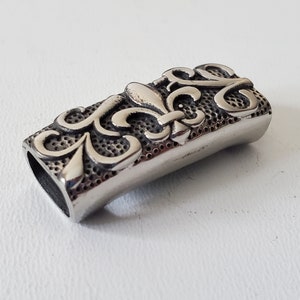 May include: A silver metal charm with a fleur-de-lis design. The charm has a textured surface and a hole for attaching to a bracelet or necklace.