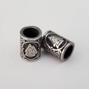 May include: Two silver metal beads with a raised design of a Celtic knot and a triangular symbol. The beads are cylindrical and have a smooth, polished finish.