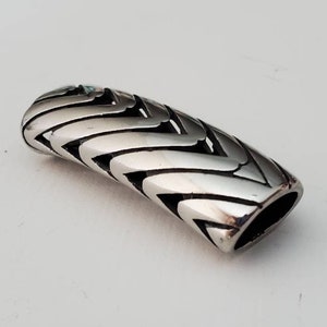 May include: A silver-colored, cylindrical bead with a woven design. The bead has a rounded top and a hollowed-out bottom. The woven pattern creates a series of open spaces along the sides of the bead. The bead is made of a shiny metal.