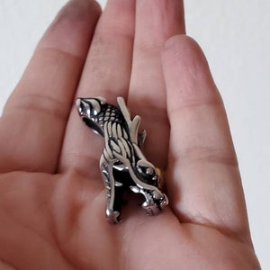 May include: A silver-colored ring featuring a detailed dragon design. The dragon is depicted in a stylized, three-dimensional form, with intricate scales and sharp features. The ring is held in a hand against a neutral background.