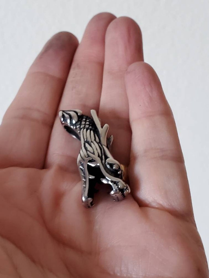 May include: A silver-colored ring featuring a detailed dragon design. The dragon is depicted in a stylized, three-dimensional form, with intricate scales and sharp features. The ring is held in a hand against a neutral background.