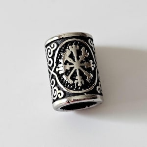 May include: A cylindrical silver-colored bead with a black center. The bead features a detailed, symmetrical design of a cross-like symbol within a circular frame, surrounded by ornate scrollwork. The bead is likely for jewelry making.