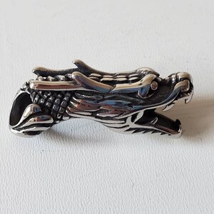 May include: A detailed, silver-colored dragon head pendant. The dragon's head is sculpted with intricate scales, sharp teeth, and a detailed mane. The pendant is designed with a hole for a chain or cord, suitable for jewelry.