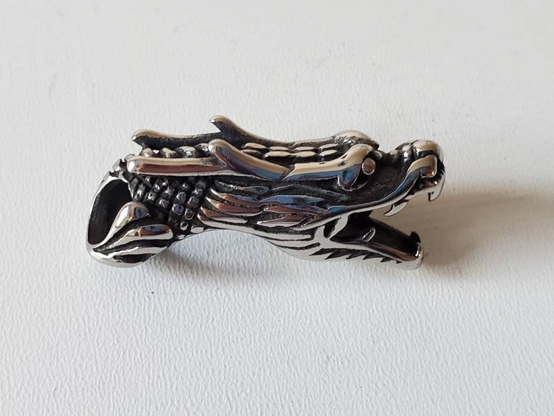 May include: A detailed, silver-colored dragon head pendant. The dragon's head is sculpted with intricate scales, sharp teeth, and a detailed mane. The pendant is designed with a hole for a chain or cord, suitable for jewelry.