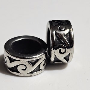 May include: Two silver-colored metal beads with a decorative, swirling pattern. The beads are cylindrical with a large central hole. The design features a series of curved lines and shapes, creating an ornate look. The beads are likely used for jewelry making or other crafts.