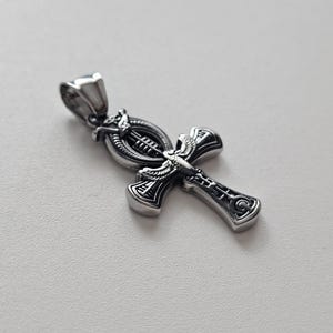 May include: A silver-toned metal ankh pendant featuring intricate engraved details, including a winged scarab motif at the center and a loop bail at the top. The surface has a polished finish with darkened recessed areas to highlight the design.