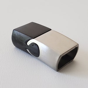 May include: A two-tone magnetic clasp with a black, rectangular top and a brushed silver-tone body. The clasp has a curved design on one end and a rectangular opening on the other.