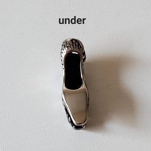 May include: A silver-colored shoe-shaped pendant with a textured top and a smooth, reflective surface. The word "under" is printed above the pendant in black letters. The pendant has a black interior.
