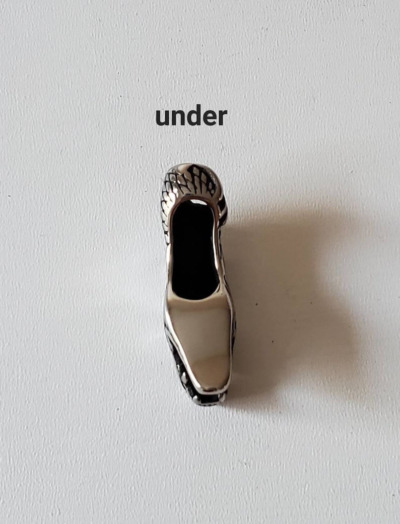 May include: A silver-colored shoe-shaped pendant with a textured top and a smooth, reflective surface. The word "under" is printed above the pendant in black letters. The pendant has a black interior.