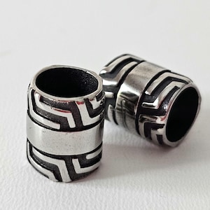 May include: Two stainless steel beads with a geometric design. The beads are cylindrical with a central band and etched patterns. One bead is upright, and the other is lying on its side.