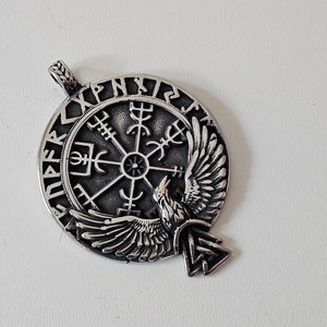 May include: A silver pendant featuring a raven with outstretched wings, a compass rose, and the inscription "Vegvisir" around the edge.