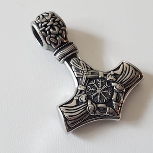 May include: A silver-colored pendant in the shape of Thor's hammer, Mjolnir. The hammer features intricate knotwork and a circular symbol. The top has a loop for a chain. The pendant is likely made of stainless steel.