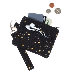 May include: A black zippered pouch with a gold star pattern and a wrist strap. The pouch is open and contains a credit card, cash, and a pair of white earbuds.