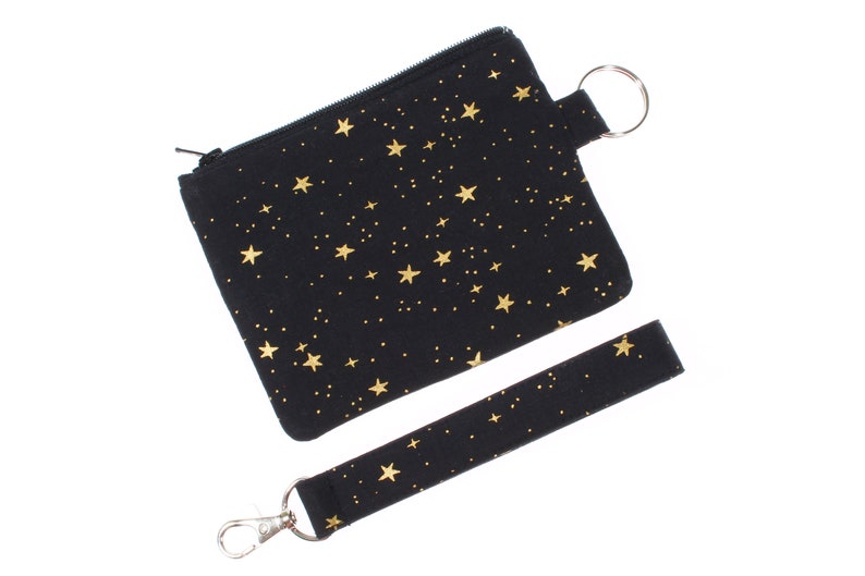 May include: A black zippered pouch with a gold star pattern and a matching black wrist strap with a gold star pattern. The pouch has a silver zipper and a silver key ring.