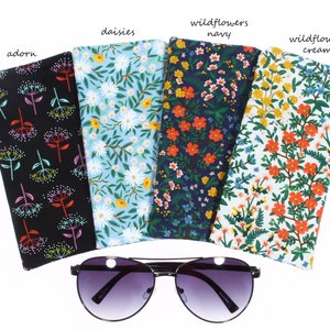 May include: Five different patterned fabric eyeglass cases, each with a floral design. The cases are arranged in a row, with the sunglasses in front of them. The cases are labeled with the names of the patterns: "adorn", "daisies", "wildflowers navy", "wildflowers cream".