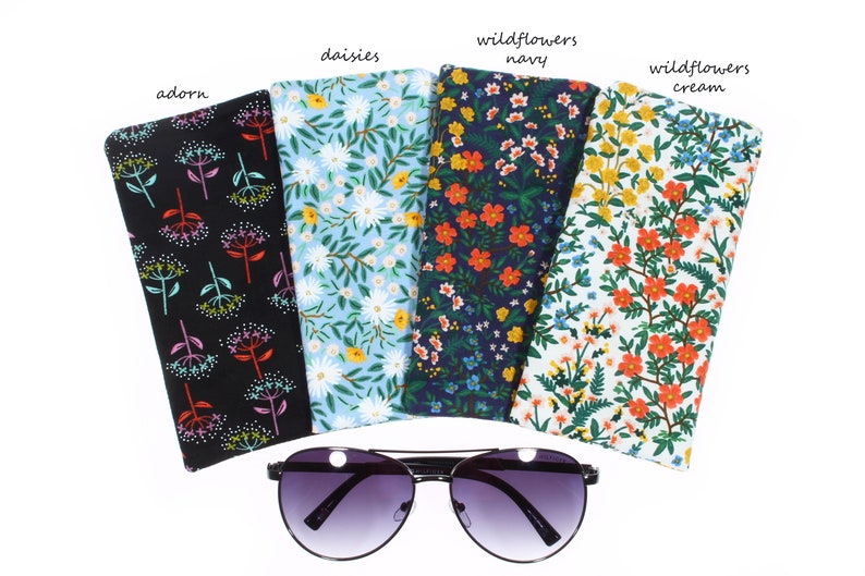 May include: Five different patterned fabric eyeglass cases, each with a floral design. The cases are arranged in a row, with the sunglasses in front of them. The cases are labeled with the names of the patterns: "adorn", "daisies", "wildflowers navy", "wildflowers cream".