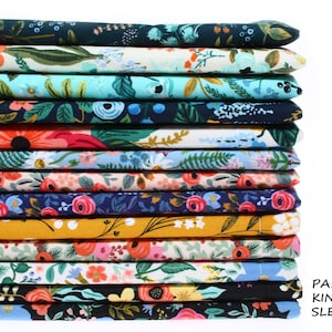 May include: A stack of 14 different floral patterned fabric squares in various colors, including red, blue, green, yellow, and white. The fabric is arranged in a stack, with the top square visible and the bottom square partially visible. The text "PADDED KINDLE SLEEVE" is visible in the lower right corner of the image.
