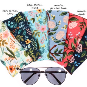 May include: Assortment of fabric eyeglass cases with floral patterns. The cases are in various colors, including cream, navy, mint, powder blue, pink, and navy. The text on the cases reads "herb garden" and "peonies".