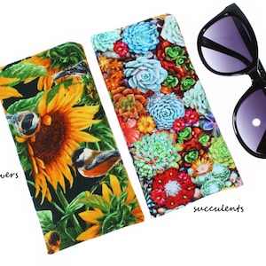 May include: Two fabric eyeglass cases with different floral patterns. One case features a sunflower design with birds, and the other case features a succulent design. Both cases are black with a white background.